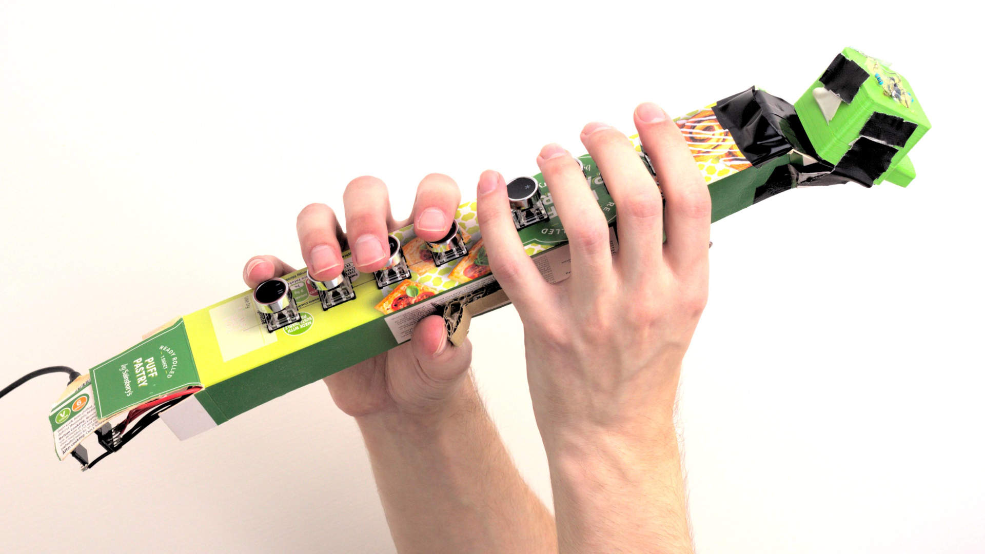 Cardboard electro-sax prototype being held up triumphantly. It's a long triangular prism shape with the 3D printed mouthpiece at one end and 8 keys down the length of the front.